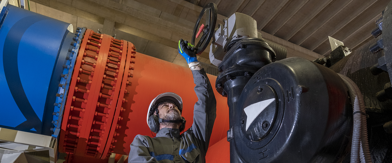 A worker turns a handle of the infrastructure. Behind him is a large orange and blue pipe