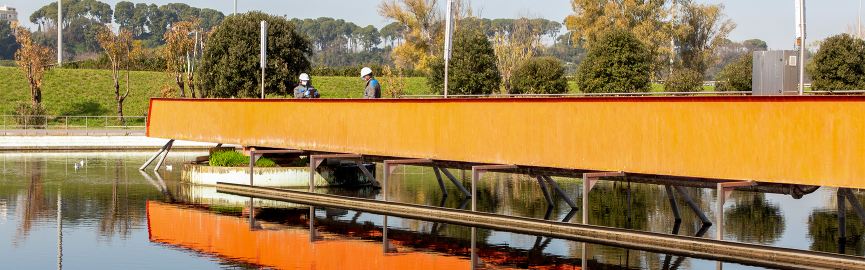 una vasca della sorgente del depuratore di roma nord con un ponte arancione in ferro su cui lavorano degli operai  acea infrastructure e gli alberi sullo sfondo