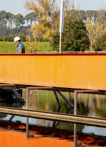  A tank at the source of the Rome North water treatment plant with an orange iron bridge on which workers are working and trees in the background.